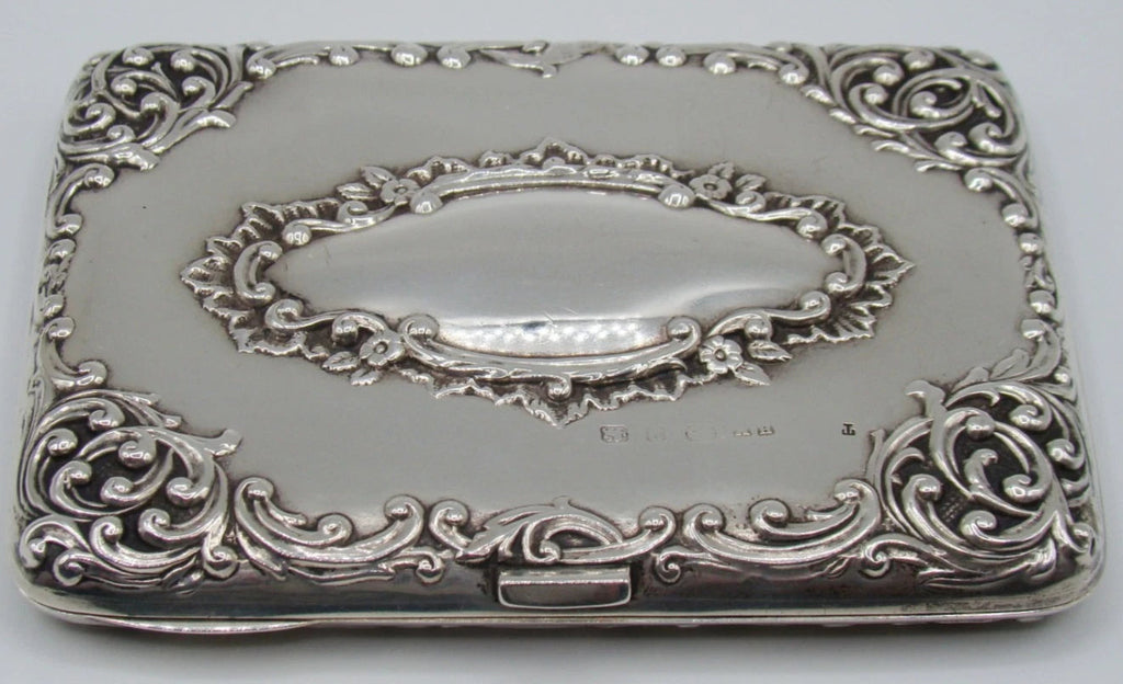 Antique silver cigarette case with raised scrolling decoration and an oval cartouche, viewed from the clasp side.