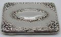 Antique silver cigarette case with raised scrolling decoration and an oval cartouche, viewed from the clasp side.