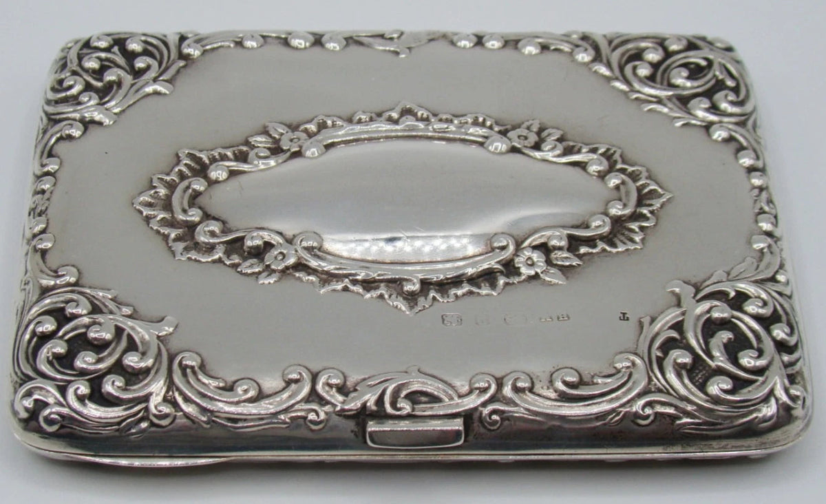 Antique silver cigarette case with raised scrolling decoration and an oval cartouche, viewed from the clasp side.