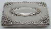 Silver cigarette case viewed from the hinge side, showing raised floral scrollwork and oval cartouche.