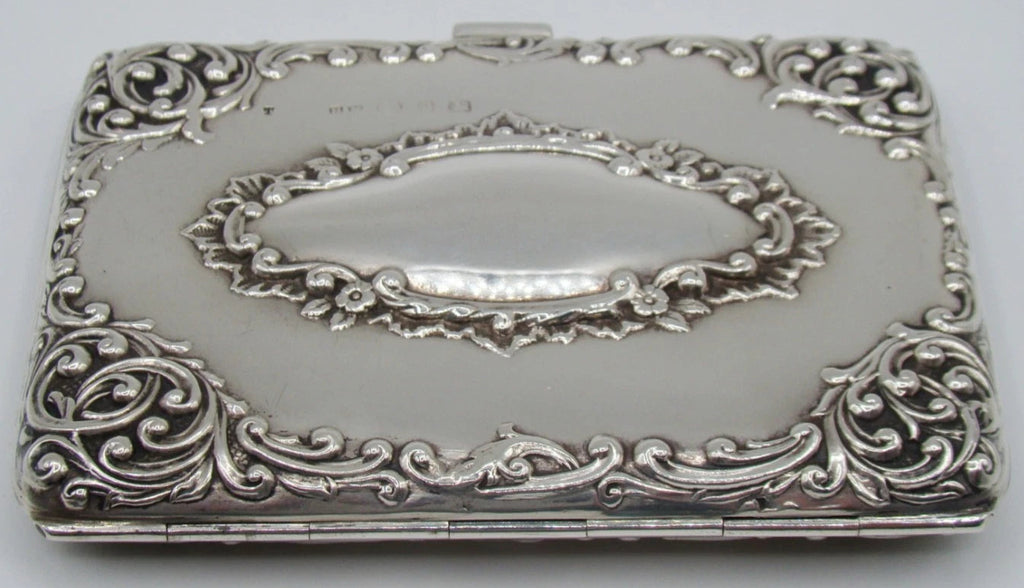 Silver cigarette case viewed from the hinge side, showing raised floral scrollwork and oval cartouche.