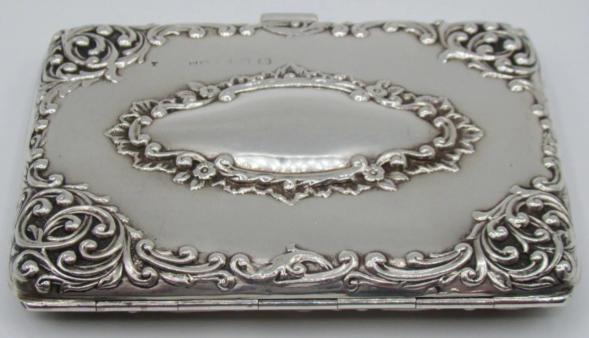 Silver cigarette case viewed from the hinge side, showing raised floral scrollwork and oval cartouche.
