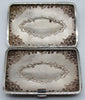 Open cigarette case showing the interior relief of the scrollwork and cartouches, with tarnish and spotting.