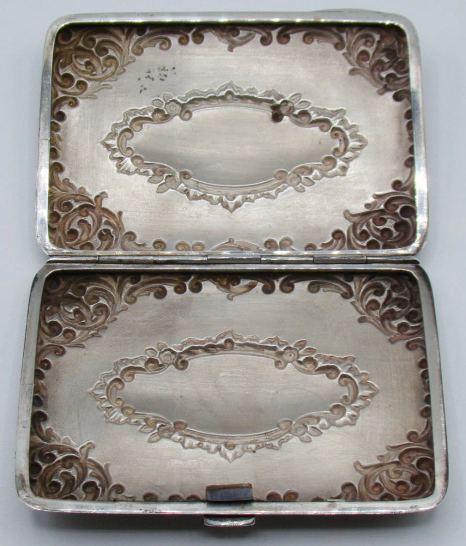 Open cigarette case showing the interior relief of the scrollwork and cartouches, with tarnish and spotting.