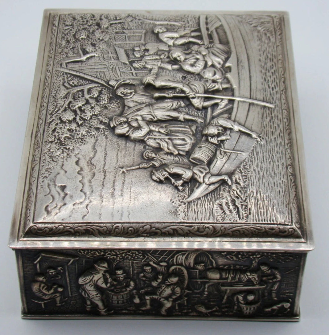 Angled view of the silver box showing the scrolling lid border and figurative side panel.