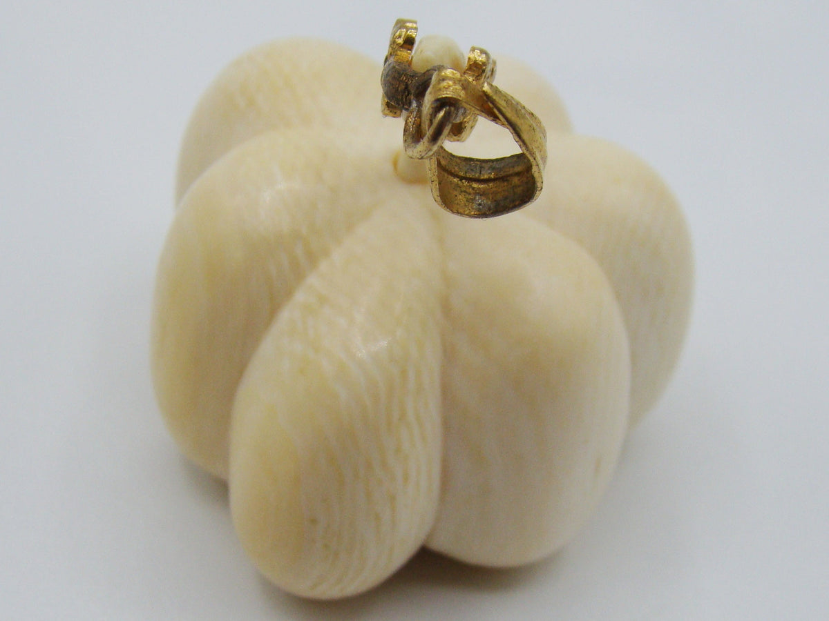 A small carved pumpkin-shaped pendant or charm in a warm ivory tone with a gold-tone loop at the top, photographed on a plain light background.